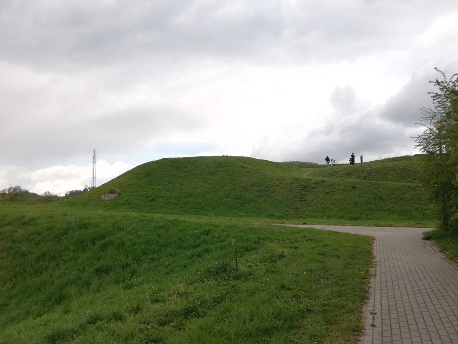 Gdańsk. St Gertrude Bastion. Contemporary photographs. The photograph presents a hill covered with grass only. There is a path trodden to the top of the hill. There are some people visible on the hill. At the bottom of the hill, there is a grey cobbled pavement. 