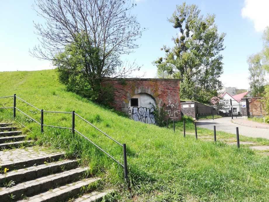 Gdańsk. St Gertrude Bastion. Contemporary photographs. The photograph presents a casemate, a shelter in the bastion stronghold – a small brick building with an arched gate. The gate is closed with the metal door of two leaves (right and left). On the left to the shelter there is a small hill covered with grass; on the right – there is a cobbled road and a private property entrance. 